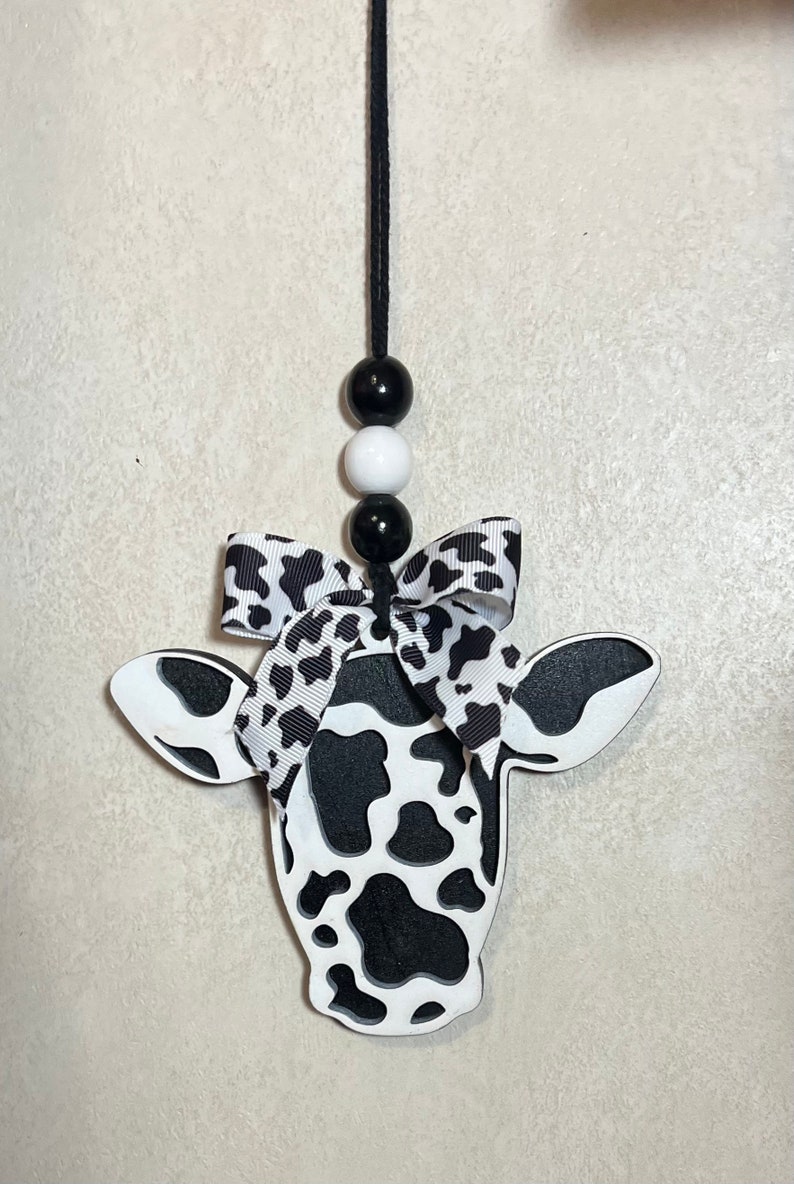 Puede incluir: Adorno con forma de vaca con dise&ntilde;o de manchas blancas y negras. Tiene una cuerda negra para colgar, con tres cuentas en blanco y negro. Un lazo con estampado blanco y negro est&aacute; atado en la parte superior de la cabeza de la vaca.