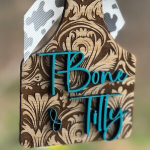 May include: A wooden cow tag with a brown floral design and the name "Bone & Tilly" in teal lettering. It is adorned with a black and white cow print ribbon and wooden beads.