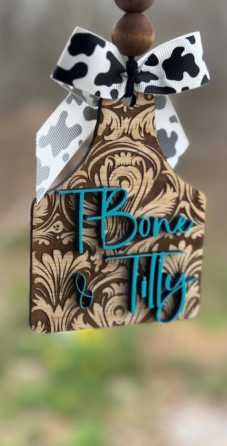 May include: A wooden cow tag with a brown floral design and the name "Bone & Tilly" in teal lettering. It is adorned with a black and white cow print ribbon and wooden beads.