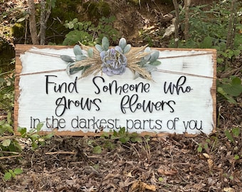 May include: White wooden sign with the text "Find someone who grows flowers in the darkest parts of you". The sign is decorated with a floral arrangement of dried flowers and greenery.