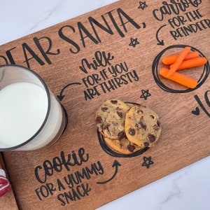 May include: A wooden tray with the words "Dear Santa, Carrots for the Reindeer" and "Milk for if you are thirsty" written on it. There is a glass of milk, a plate of cookies, and a plate of carrots on the tray. The tray is decorated with stars and hearts.