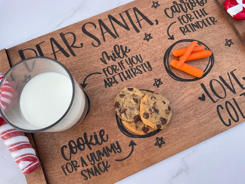May include: A wooden tray with the words "Dear Santa, Carrots for the Reindeer" and "Milk for if you are thirsty" written on it. There is a glass of milk, a plate of cookies, and a plate of carrots on the tray. The tray is decorated with stars and hearts.