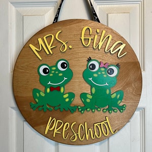 May include: A round wooden sign with the words "MRS. Gina PRESCHOOL" in yellow lettering. Two green frog illustrations are featured below the name. The sign is hanging by a black and white ribbon.