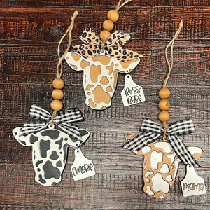 May include: Three wooden cow ornaments with black and white or brown and white spots. Each ornament has a tag with a different name: "Boss Babe", "Emere", and "Mama".