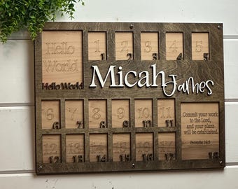 Puede incluir: Un tablero de hitos de madera con el nombre "Micah James" en blanco. Tiene espacios para fotos y los números 1-18. Las palabras "Hello World" están en la parte superior izquierda. Un versículo bíblico está en la parte inferior derecha.