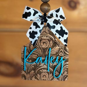 May include: Wooden tag with a floral design and the name "Kailey" in turquoise script. The tag is topped with a black and white cow print bow and wooden beads, hanging from a black string. The tag is a decorative item.