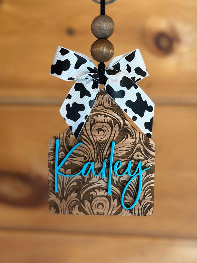 May include: Wooden tag with a floral design and the name "Kailey" in turquoise script. The tag is topped with a black and white cow print bow and wooden beads, hanging from a black string. The tag is a decorative item.