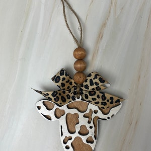 Puede incluir: Adorno en forma de vaca con centro de vetas de madera y contorno plateado. El adorno tiene un lazo con estampado de leopardo y tres cuentas de madera. Se adjunta una cuerda de yute para colgar.