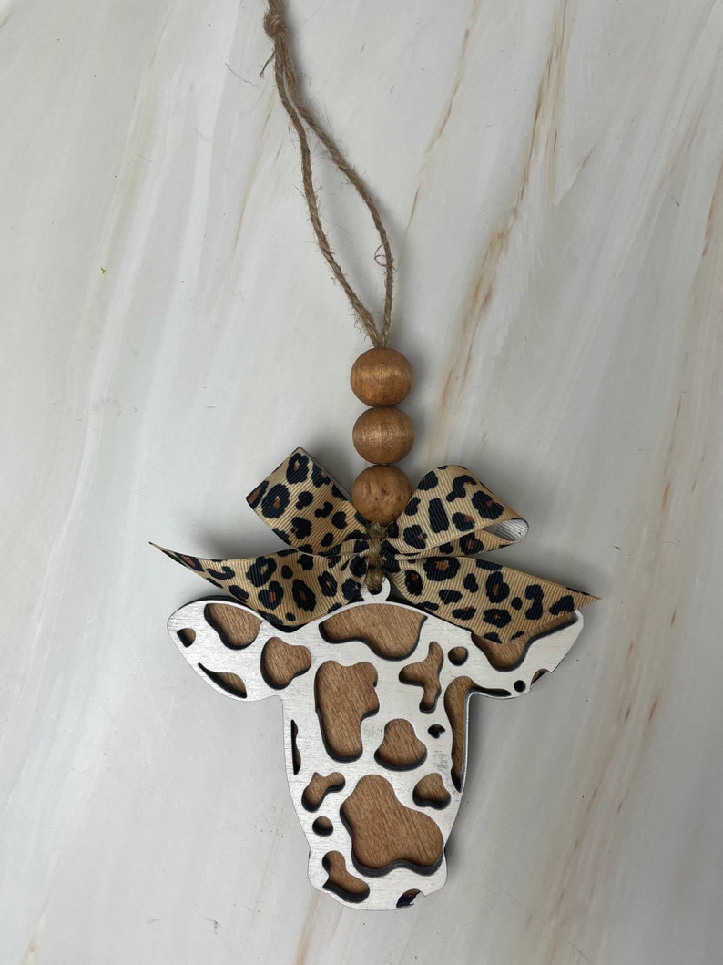 Puede incluir: Adorno en forma de vaca con centro de vetas de madera y contorno plateado. El adorno tiene un lazo con estampado de leopardo y tres cuentas de madera. Se adjunta una cuerda de yute para colgar.