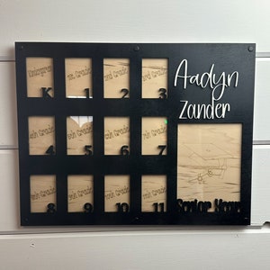 May include: Black wooden photo display with spaces for 12 photos, labeled "Kindergarten" through "11th Grade." The name "Aadyn Zander" is displayed in white script, with a larger space for a senior year photo. The words "Senior Year" are also displayed.