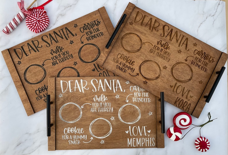May include: Three wooden trays with black handles, each with the text "Dear Santa" and a space for milk, cookies, and carrots for the reindeer. The trays are decorated with stars and hearts. The text "Love" is written on each tray, followed by a name.