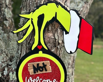 Op de afbeelding: Een groene Grinch-hand die een rode en zwarte kerstbal vasthoudt met de woorden "Not Welcome" erop.