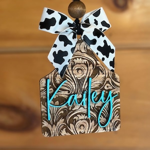 May include: A wooden ornament with a brown floral design and the name "Kailey" in turquoise script. It is topped with a black and white cow print bow and wooden beads. The ornament hangs from a black string.