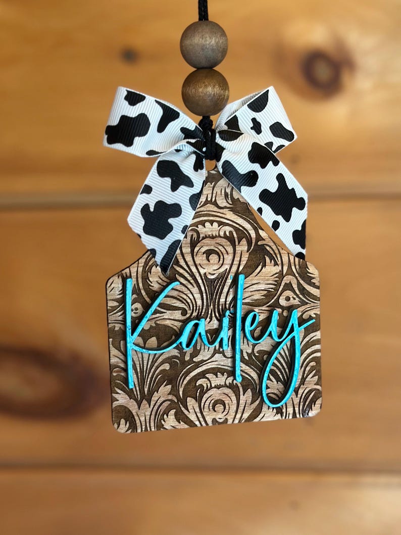 May include: A wooden ornament with a brown floral design and the name "Kailey" in turquoise script. It is topped with a black and white cow print bow and wooden beads. The ornament hangs from a black string.