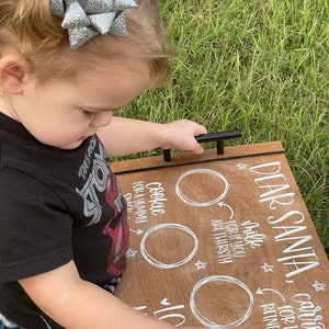May include: A wooden tray with white painted text that reads "Dear Santa, carrots for the reindeer, cookie for a yummy snack, milk for if you are thirsty". The tray has two black metal handles and three white circles for the items to be placed.