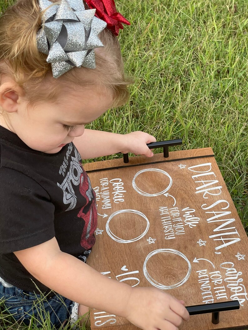 May include: A wooden tray with white painted text that reads "Dear Santa, carrots for the reindeer, cookie for a yummy snack, milk for if you are thirsty". The tray has two black metal handles and three white circles for the items to be placed.