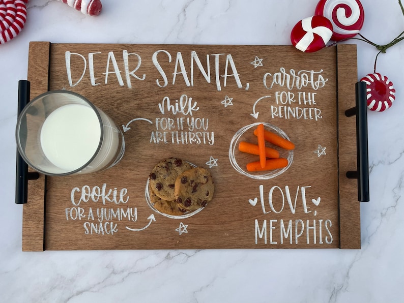 May include: A wooden tray with the words "Dear Santa, carrots for the reindeer, milk for if you are thirsty, cookie for a yummy snack, love, Memphis" written in white paint. The tray contains a glass of milk, two chocolate chip cookies, and four baby carrots.