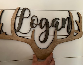Puede incluir: Cuernos de ciervo de madera con el nombre "Logan" recortado en el centro. Los cuernos están teñidos de un color marrón claro y el nombre está teñido de un color marrón oscuro.