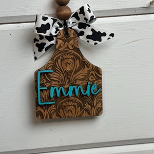 May include: A wooden tag with the name "Emmie" in turquoise lettering. The tag has a brown, ornate design and is topped with a black and white cow print bow and wooden beads. The tag hangs from a black string.