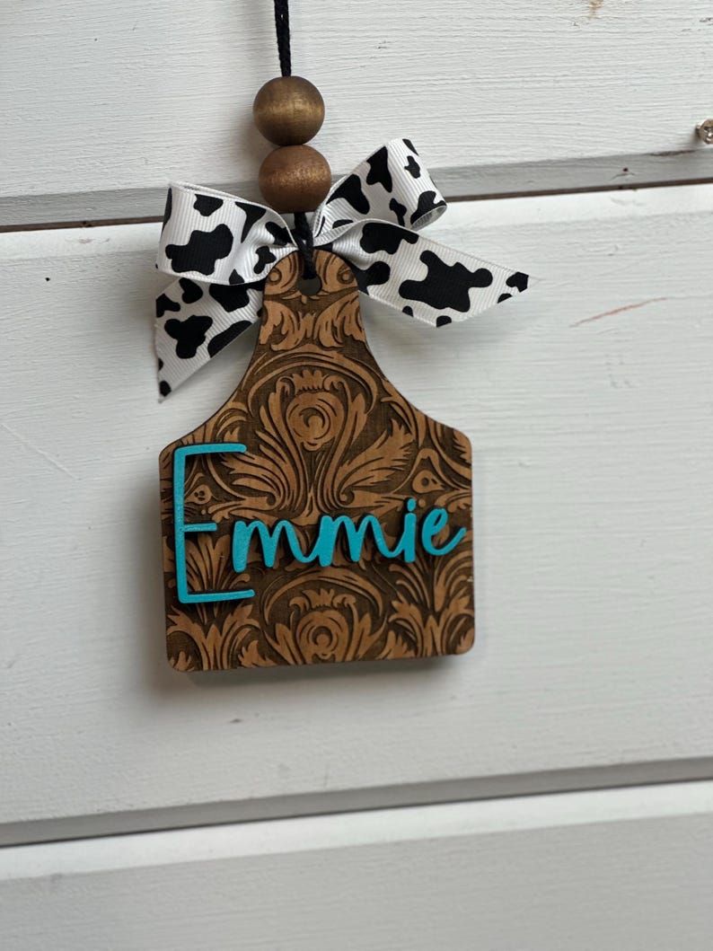 May include: A wooden tag with the name "Emmie" in turquoise lettering. The tag has a brown, ornate design and is topped with a black and white cow print bow and wooden beads. The tag hangs from a black string.