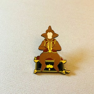 Op de afbeelding: Een bruine emaille pin met een aapachtig figuur dat op een decoratieve kist zit. Het figuur draagt een rood vest over een geel overhemd. De kist heeft een zwart-gouden ontwerp.