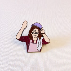 Puede incluir: Un pin de esmalte morado con un personaje de dibujos animados con cabello castaño que lleva un sombrero morado y un delantal rosa. La cara del personaje está cubierta por un círculo con el texto "I'm Breaking Down".