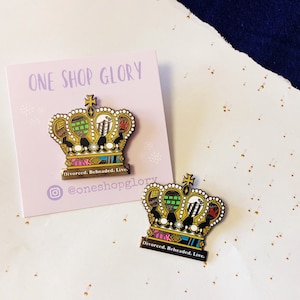 May include: Two gold-colored enamel pins shaped like crowns with colorful designs and the text "Divorced. Beheaded. Live."  The pins are on a white surface with gold glitter.