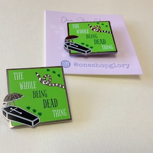 May include: Green enamel pin with a black coffin, a green snake, and a black umbrella. The text reads "The Whole Being Dead Thing".