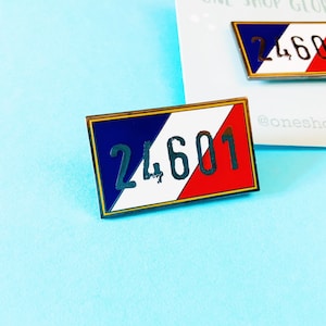 Puede incluir: Un pin de metal con un diseño azul, blanco y rojo, que se asemeja a la bandera francesa. El número "24601" está impreso en negro en la franja blanca.