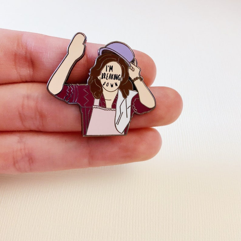 May include: A purple enamel pin featuring a cartoon character with brown hair wearing a purple hat and a pink apron. The character's face is obscured by the text "I'M BREAKING DOWN".