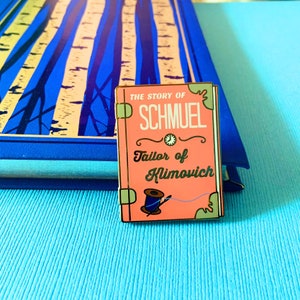 Op de afbeelding: Een roze emaille pin met gouden accenten die een boekomslag voorstelt. De titel van het boek is "The Story of Schmuel Tailor of Klimovich".