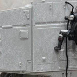May include: A gray plastic model of an AT-AT walker featuring a small, detailed figure of a character in silver armor and a black cape climbing the side of the vehicle using a black rope. The walker has mechanical paneling and a jointed neck section.
