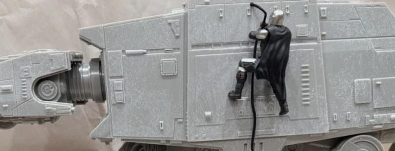 May include: A gray plastic model of an AT-AT walker featuring a small, detailed figure of a character in silver armor and a black cape climbing the side of the vehicle using a black rope. The walker has mechanical paneling and a jointed neck section.