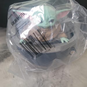 May include: A green, large-eared creature figurine with dark eyes, wearing a tan robe, seated inside a gray, bowl-shaped hover pod. The item is enclosed in a clear plastic bag featuring a barcode, the text Golden Link, and Made in China.