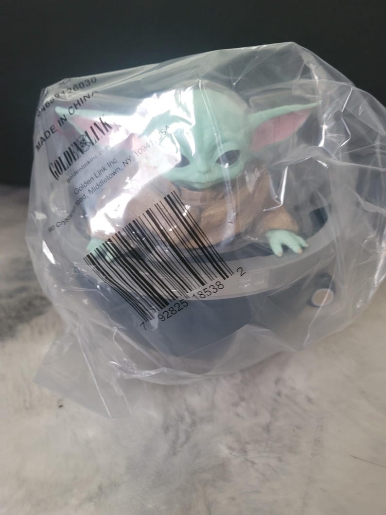 May include: A green, large-eared creature figurine with dark eyes, wearing a tan robe, seated inside a gray, bowl-shaped hover pod. The item is enclosed in a clear plastic bag featuring a barcode, the text Golden Link, and Made in China.