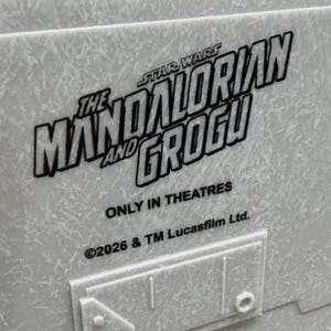 May include: A light gray textured surface featuring the black printed text THE MANDALORIAN AND GROGU, STAR WARS, ONLY IN THEATRES, and 2026 & TM Lucasfilm Ltd. Below the text is a raised rectangular panel with a small square detail on the right.