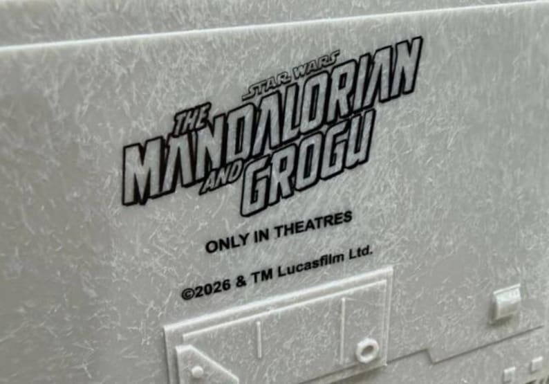 May include: A light gray textured surface featuring the black printed text THE MANDALORIAN AND GROGU, STAR WARS, ONLY IN THEATRES, and 2026 & TM Lucasfilm Ltd. Below the text is a raised rectangular panel with a small square detail on the right.
