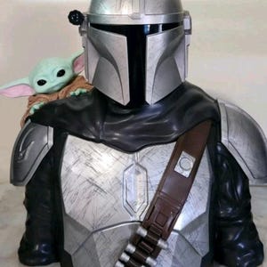 K&ouml;nnte beinhalten: Eine B&uuml;ste einer silbernen R&uuml;stungsfigur mit Helm und schwarzem Visier, die ein verwittertes Metallic-Finish aufweist. Eine kleine gr&uuml;ne Kreatur mit gro&szlig;en Ohren und einem beigen Gewand sitzt auf der Schulter der gepanzerten Figur.