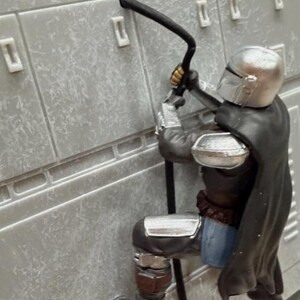 May include: A plastic action figure of a character wearing a metallic silver helmet and armor with a black cape. The figure is positioned against a light gray textured wall panel with rectangular indentations, holding a black curved cable.