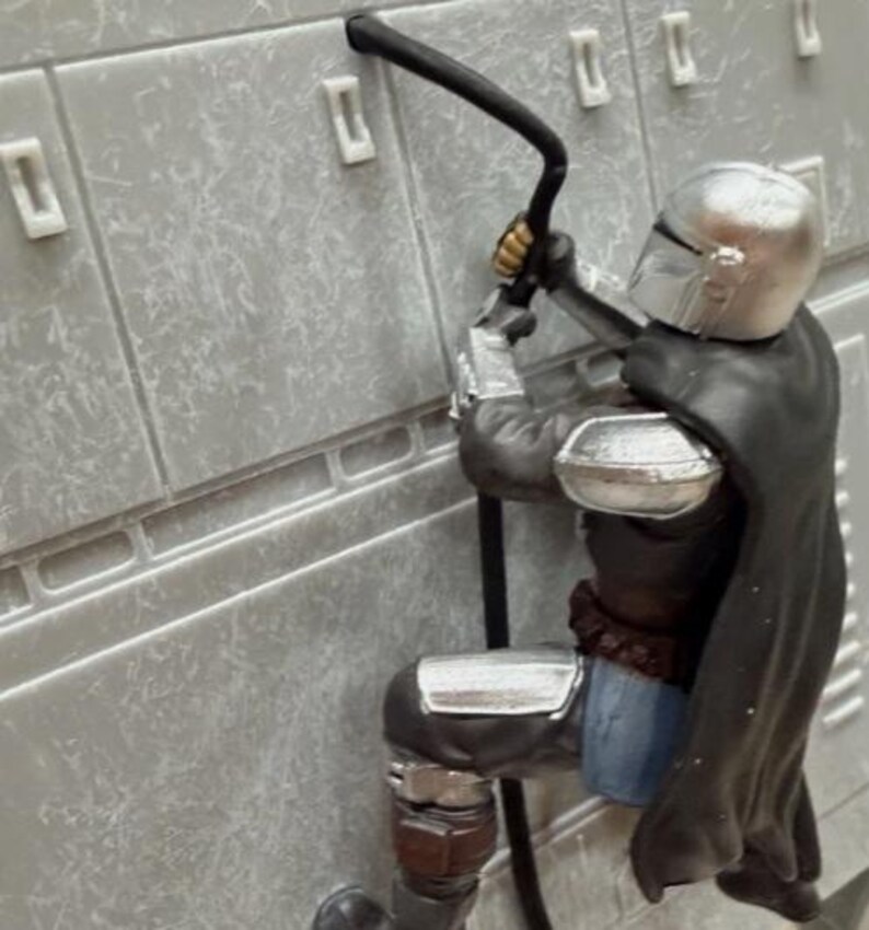 May include: A plastic action figure of a character wearing a metallic silver helmet and armor with a black cape. The figure is positioned against a light gray textured wall panel with rectangular indentations, holding a black curved cable.