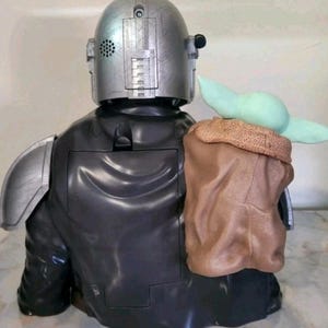 K&ouml;nnte beinhalten: Eine Kunststofffigur eines gepanzerten Charakters mit silbernem Helm und schwarzem Anzug, der eine kleine gr&uuml;ne Kreatur mit gro&szlig;en Ohren in einer strukturierten braunen Tasche tr&auml;gt, die am R&uuml;cken der Schulter und des Torsos befestigt ist.