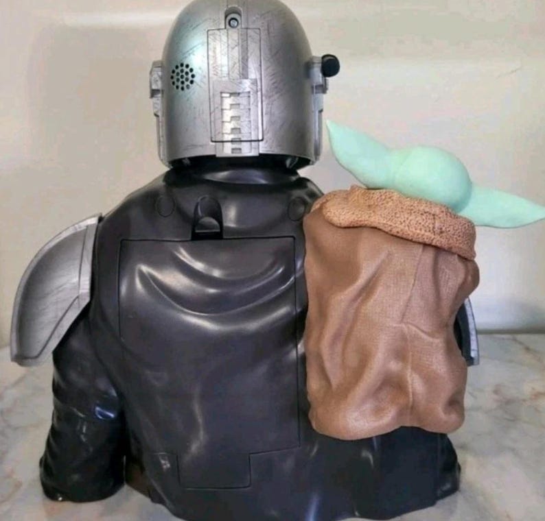 K&ouml;nnte beinhalten: Eine Kunststofffigur eines gepanzerten Charakters mit silbernem Helm und schwarzem Anzug, der eine kleine gr&uuml;ne Kreatur mit gro&szlig;en Ohren in einer strukturierten braunen Tasche tr&auml;gt, die am R&uuml;cken der Schulter und des Torsos befestigt ist.