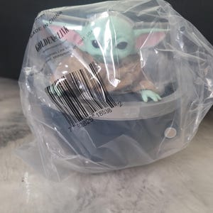 May include: A small green creature with large ears and eyes, wearing a tan robe, sits inside a dark gray circular pod. The figure is encased in clear plastic packaging featuring a barcode, the text Golden Link, and Made in China.