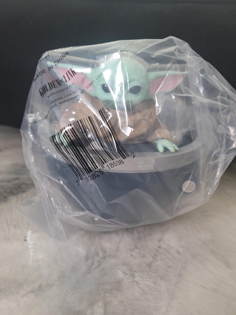May include: A small green creature with large ears and eyes, wearing a tan robe, sits inside a dark gray circular pod. The figure is encased in clear plastic packaging featuring a barcode, the text Golden Link, and Made in China.
