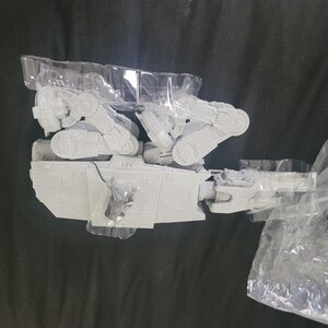 May include: A light gray plastic model of an AT-ST walker vehicle from Star Wars, partially encased in a clear molded plastic tray. The model features articulated legs and a detailed cockpit section. A plastic bag with the text Mandalorian is visible.