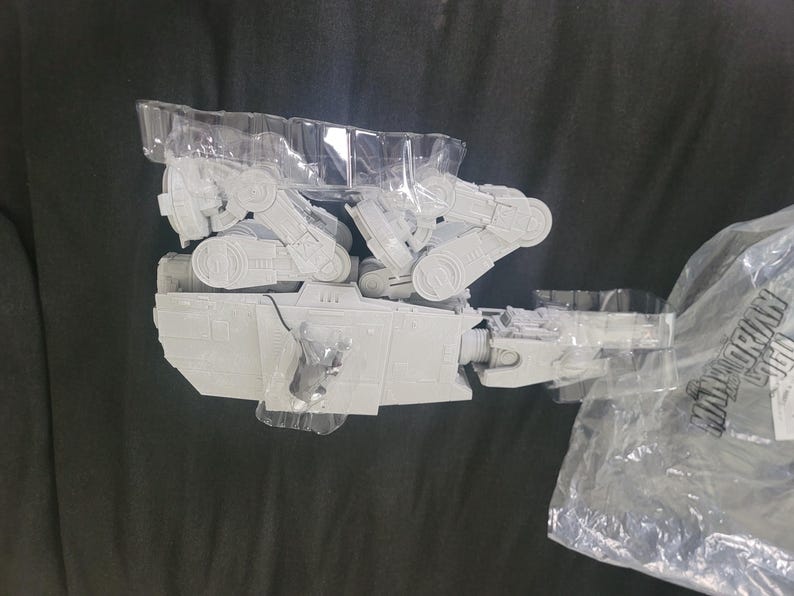 May include: A light gray plastic model of an AT-ST walker vehicle from Star Wars, partially encased in a clear molded plastic tray. The model features articulated legs and a detailed cockpit section. A plastic bag with the text Mandalorian is visible.