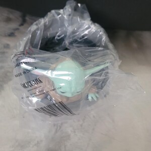 May include: A small green and tan creature figurine with large ears and eyes, encased in a clear plastic bag. The packaging features printed text including "MADE IN CHINA", "GOLDEN LINK", and a barcode, partially obscuring the figure inside.