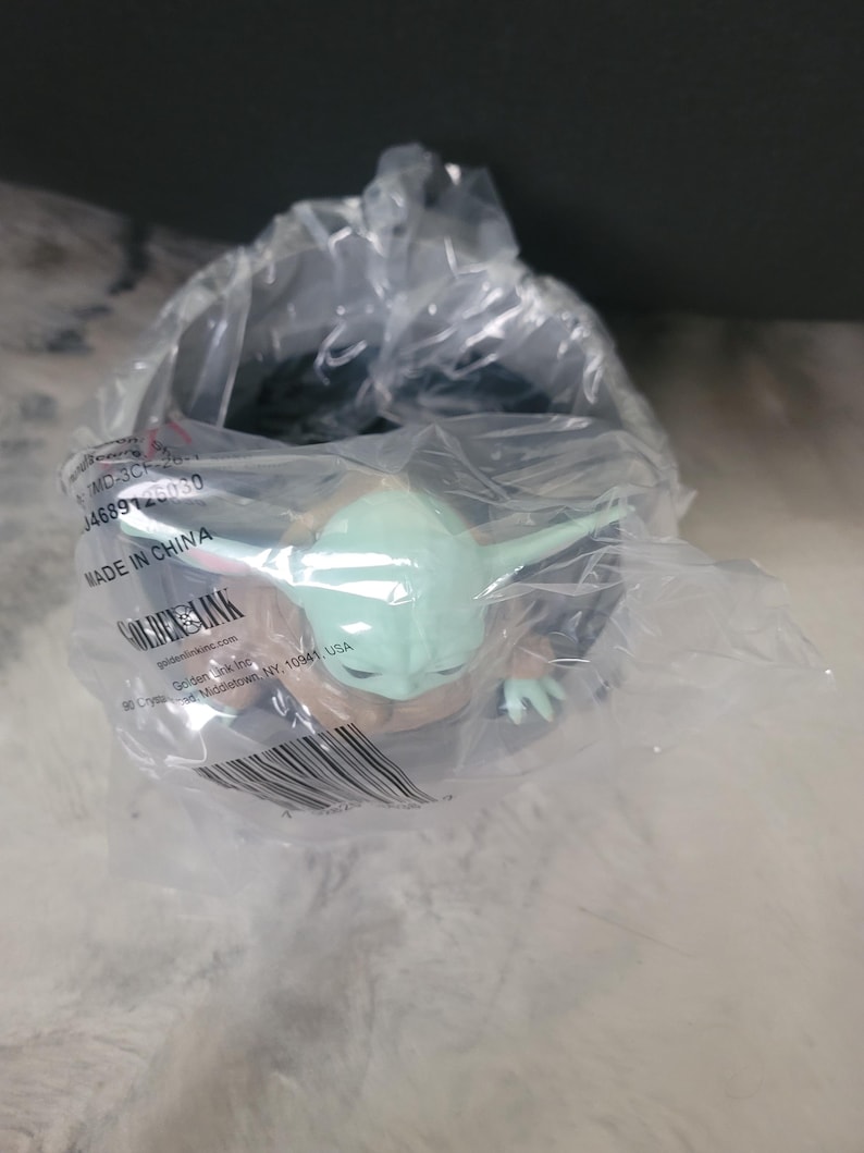 May include: A small green and tan creature figurine with large ears and eyes, encased in a clear plastic bag. The packaging features printed text including "MADE IN CHINA", "GOLDEN LINK", and a barcode, partially obscuring the figure inside.