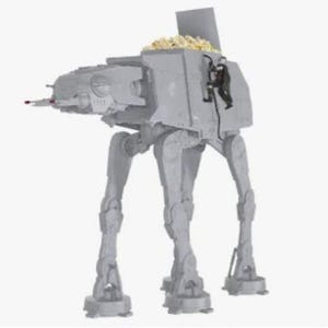 May include: A gray plastic AT-AT walker popcorn bowl featuring a hinged top hatch that is open, revealing a compartment filled with popcorn. A small black action figure is attached to the side of the walker, appearing to climb the exterior.