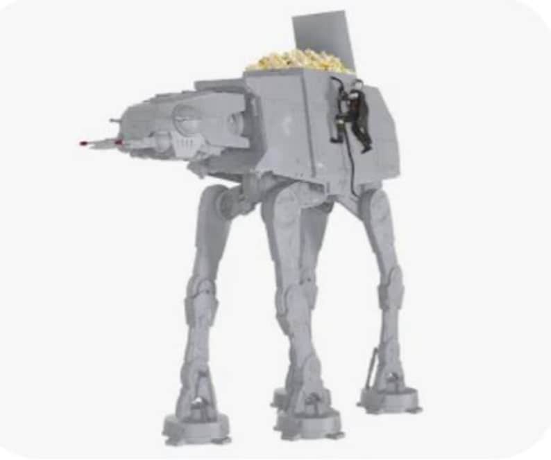May include: A gray plastic AT-AT walker popcorn bowl featuring a hinged top hatch that is open, revealing a compartment filled with popcorn. A small black action figure is attached to the side of the walker, appearing to climb the exterior.